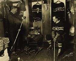 two men, one in suit, one in hockey uniform, look at the hockey equipment of a player in the dressing room.