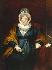 Oil painting of a woman sitting in a striped chair. She is wearing a dark-colored dress, with a shawl, contrasted with her tight, white cap and collar. Next to her is a table with writing instruments.