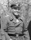 A smiling man wearing a military uniform, peaked cap with skull emblem and neck order in the shape of a cross.