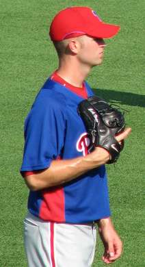 A man in a blue baseball jersey, gray baseball pants, and a red baseball cap stands on a green field. He has a black baseball glove on his right hand.