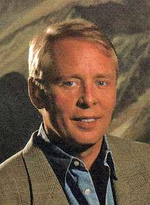 A blond, blue-eyed man wearing a brown sport jacket and open-necked collared blue shirt