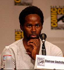 A black man wearing a white shirt. A panel with the logo for the San Diego Comic-Con is seen behind him. In front of him are a water bottle, a plastic cup, a microphone and a paper reading "Damon Lindelof".