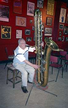 An elderly, grey-haired man sits in front of a wall covered with framed photographs and posters, and plays a floor-mounted contrabass saxophone.