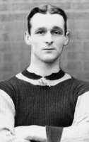 Clean-shaven white man with neatly trimmed hair wearing a dark-coloured knitted sports jersey with light sleeves and collar trim