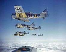 Silver fighter aircraft, marked with the British Royal Air Force's distinctive roundel, fly above the clouds in formation