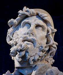 Stone image of the head of a man, showing signs of erosion due to age. The most striking feature is the heavily coiled beard and hair. The eyes are looking sightlessly upwards