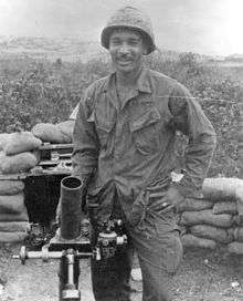 A black and white image showing Hector from the waist up in his military combat uniform with kevlar helmet. His hands are on his hips and he is smiling at the camera.