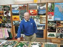 Heinz Stücke stands in front of an array of photos pinned to corkboard. He smiles slightly and leans against one of several glass-covered trays of trinkets on a table before him.