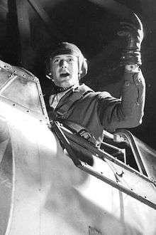 A pilot seated in his fighter craft gestures with a gloved hand.