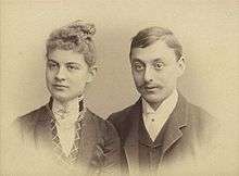 A charming and iconic period photo of a young couple around 1900. The lady has her hair swept upwards and is wearing a high collar fastened with a jewel. The gentleman sports a gallant moustache neatly trimmed and is also wearing a high collar.