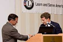 a Native American Hispanic man dressed in a brown suit shakes hands with a younger man with curly hair wearing a dark blue suit. Behind the two men is a Learn and Serve American banner.