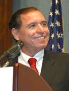 Head and shoulders of a man in his forties with close-cropped dark hair, dressed in a dark blue suit, a white shirt and a red striped tie. On his right lapel is the President’s "Call to Service" Award medal. Over his right shoulder the U.S. flag.