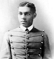 Cadet Henry O. Flipper in his West Point cadet uniform. It has three large round brass buttons left, middle and right showing five rows. The buttons are interconnected left to right and vice versa by decorative thread. He is wearing a starched white collar and no tie. He is a lighter colored African-American with plated corn rows of neatly done hair. He is facing the camera and looking to the left of the viewer.