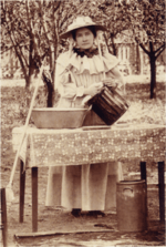 Sepia-toned photograph of a woman in a bonnet by a table outdoors.