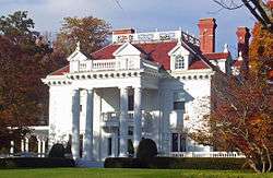 A white house with a colonnaded front and dormer windows in its red roof with brick chimneys on top seen from slightly to the right of center. The sun lights the house from the left and on either side are trees showing fall color