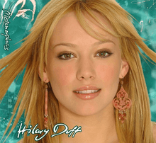 The face of a young blonde girl standing in front of a green background. She has brown eyes and is wearing brown-and-pink chandelier earrings. On her image, the words "Hilary Duff" and "Metamorphosis" are written in white, cursive print.
