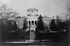 A black-and-white photo of a building situated on a hill and partially obscured by trees. A cupola with a clock sits on the roof.