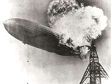 A black and white photograph of an airship near a mooring mast exploding at its stern.