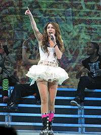 A brunette teenager singing into a microphone. She is facing up and pointing with her index finger. She is wearing a short white dress with a tutu bottom. Behind her are background dancers and graffiti projected on a screen.