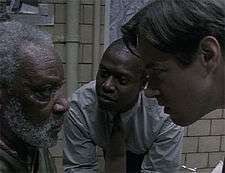 Two men lean in closely while speaking with a bearded, unhappy-looking man inside a police interrogation room.