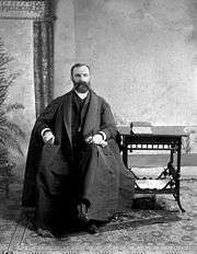 A posed formal shot of Wilson, who is seen as a young man, though with full beard and mustache. He sits next to a table on which is a book, and wears a long coat or cape which extends well around him.
