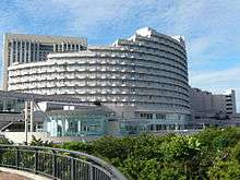 large circular multi-story building with stepped upper levels, facade consisting of multiple rows of balconies; foreground is a tree-lined park area, with a curved walkway railing, and additional buildings behind