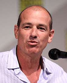 A balding man with a buttoned shirt in front of a microphone, and he is talking.