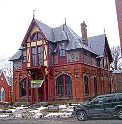 An ornate two-story building with many peaked and pointed roofs. The ground floor is brick while the upper story is wood. In front is a lawn with some snow and a green car parked in the street on the right.