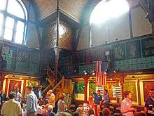 A large space with a wooden gallery along the upper portion, large windows above, and paintings and ornate decorations in the wooden walls. On the lower level, at the bottom of the image, there are a lot of people doing various things