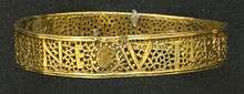A gold bracelet with a pattern and writing. The upper and lower edges are solid, but between them is a lacey pattern made of leafy plant tendrils. Amidst this mostly perforated pattern, letters are formed from solid gold segments.