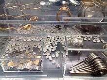Transparent perspex box containing a shelf, two inner boxes, a large silver dish and many dozens of coins. The smaller inner box contains coins, whereas the larger contains two goblets and stacked ladles. The shelf holds gold bracelets, gold chains, and engraved spoons.