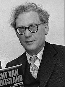 A black-and-white photograph of a grey-haired and spectacled man in his sixties is looking at the viewer. He wear a dark suit and tie, and is holding a copy of one of the books he has written.