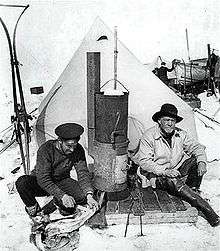 Two men sitting in front of a pointed tent; left-hand man in flat naval cap is skinning a penguin, right-hand man wears a wide-brimmed hat. Between them is a tall stove. Other equipment is visible in the background