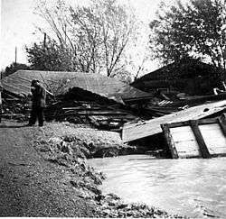 Planks of wood are scattered on the ground; a roof is visible on top of them; a man stands with his head bowed in front of the rubble.