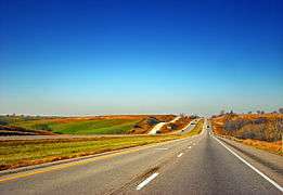 A highway underneath a clear sky surrounded by harvested cropland and green pastures.