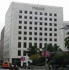 A color photograph of a ten-story retail storefront showing a plain white stone exterior containing eight rows and eight columns of windows set flush with the white stone. Above the windows, the name of the store is shown in lower-case gray letters: Macy's.