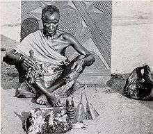 A black and white photo of an Igbo traditional spiritual practitioner known as dibia wearing a cloth alung round the should and sitting cross legged on an outside sand floor, tools of his practice are in front of him including what appear to be animal skins, a small carved image of Ikenga, and a bell. He has a white line of chalk over his eyes and is slightly bowing. In the background is a carved door with intricate lines carved into it, appearing to stick out the ground behind the man.