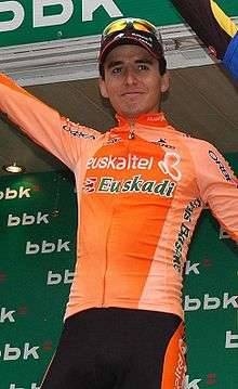 A man in his mid twenties wearing a mostly orange cycling jersey with white trim standing in front of a green background.