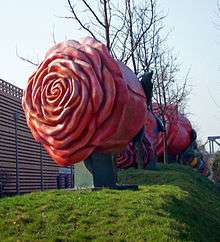 A large metallic sculpture of a red rose on a small grassy mound, with bare trees and other similar sculptures in the background