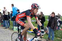 A road racing cyclist wearing a black and red jersey with white trim riding down the road. Spectators are visible on the roadside.