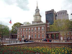The Front of Independence Hall on an overcast day. The steeple and facade of the building are visible, and an American flagpole stands on the building's right side. The foreground has a flower garden surrounded by a low brick wall with a fence on top. In the background, trees surround the building, and the Penn Mutual Tower and the Penn Mutual Life Building are visible, located to the right of the steeple relative to the viewer.