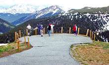 A small paved area encircled by a guardrail with wooden posts and ropes. People are standing in it looking at high snow-capped mountains in the distance