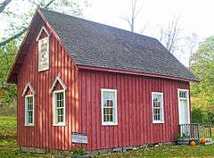 A small red wooden house with vertical siding, a black roof, white trim and a small wooden porch near the right with two pumpkins at the bottom. Signs on the lefthand side of the building and at the bottom, on the ground, both read "Indian Rock Schoolhouse"