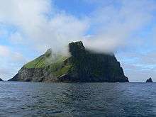 A precipitous and cliff-girt green island is mist-shrouded near its summit but with blue skies above.