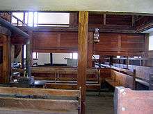 A room with wooden benches and paneled wood and plaster walls with an iron stove in the middle. An exhaust pipe leads out from the stove to the left. There is an oil lamp on a post in the middle of the picture.