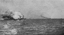 A ball of flame engulfs a large gray warship. Several smaller ships are seen in the distance.