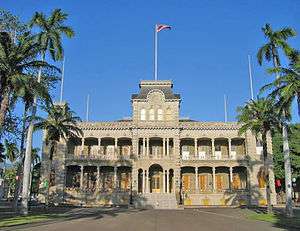 Photograph of the front of ʻIolani Palace, flanked by palm trees with the Hawaiian flag flying atop the central tower.