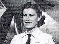 A woman in a neat, white uniform with a black tie with dark hair that is tied back, stands at attention in front of an airplane and only the area from her shoulders up are visible in black and white. She is smiling.