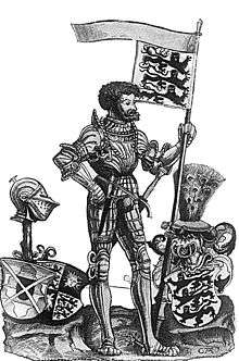 A dark haired and bearded man wearing armor and carrying a flag showing three lions. He is standing beside two three shields, two with the same symbols as his flag, and one topped by a helmet.