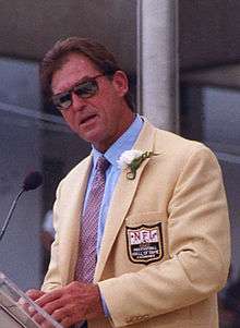 Color photo of Jack Youngblood, 51-year-old white man dressed in gold jacket, blue shirt, tie and sunglasses, giving his Pro Football Hall of Fame induction speech in 2001.